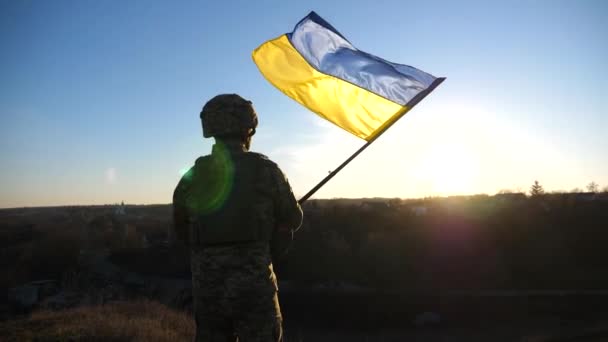 Soldier of ukrainian army stands at hill and holds waving flag of Ukraine. Man in military uniform and helmet lifted up flag against sunset. Victory against russian aggression. Invasion resistance. - Séquence, vidéo