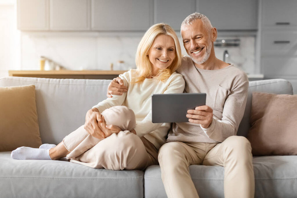Красивые, любящие старшие спаусы с помощью цифрового планшета, сидя дома на диване. Счастливые пожилые мужчина и женщина платят за товары и услуги в Интернете, с помощью банковского или торгового приложения, копировать пространство - Фото, изображение