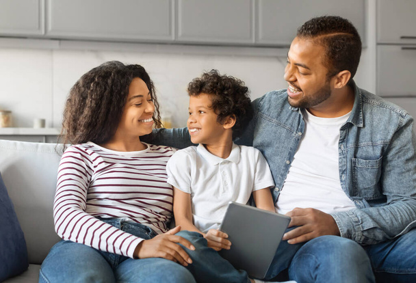 Familia afroamericana feliz con la tableta digital que relaja junta en el hogar, padres negros sonrientes y niño preadolescente que usa el gadget moderno mientras se sienta en el sofá en el interior del salón - Foto, Imagen