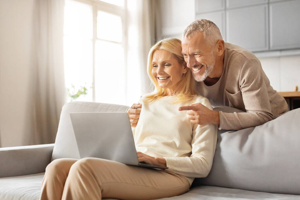 Зрелые люди и цифровой мир. Счастливая старшая пара с помощью ноутбука вместе просматривая Интернет, сидя на диване дома, скопировать пространство. Отставной муж и жена читают онлайн-новости на компьютере в выходные - Фото, изображение