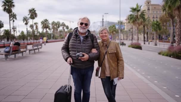 Fotoaparát zoomující usmívající se kavkazský dospělý turistický pár stojící a pózující na ulici se zavazadly. Manžel a manželka užívají své důchodce prázdniny na sobě zimní oblečení na slunný den - Záběry, video