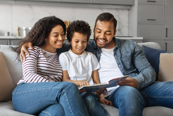 Χαρούμενη Αφρο-αμερικανική οικογένεια με το μικρό γιο ανάγνωση του βιβλίου μαζί στο σπίτι, φροντίδα μαύρο γονείς δεσμός με τους Preteen αρσενικό παιδί, χαλαρώνοντας στον καναπέ στο σαλόνι, Αντιγραφή χώρου - Φωτογραφία, εικόνα