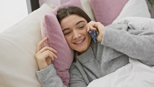 Eine junge Frau plaudert fröhlich auf einem Smartphone, während sie es sich auf einem Bett in einer gemütlichen, gut beleuchteten Schlafzimmeratmosphäre gemütlich macht. - Filmmaterial, Video