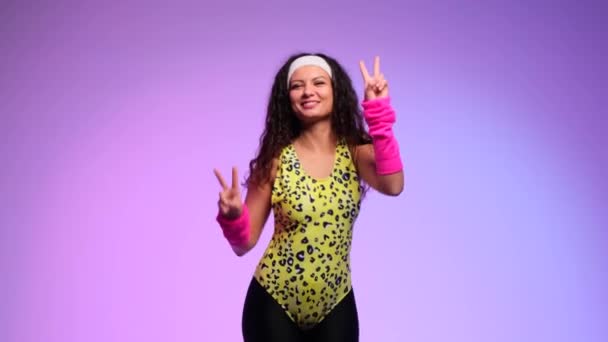 Nerdy kudrnatá žena v pestrobarevném oblečení z 80. let ukazuje vítězné gesto a tancuje s úsměvem na purpurovém pozadí. Symbolizující vítězství a radost. - Záběry, video