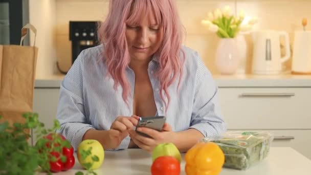 Eten online bestellen. Glimlachende blanke vrouw met telefoon kopen boodschappenbezorging van internet tijdens het koken in de keuken. Hoge kwaliteit FullHD beeldmateriaal - Video