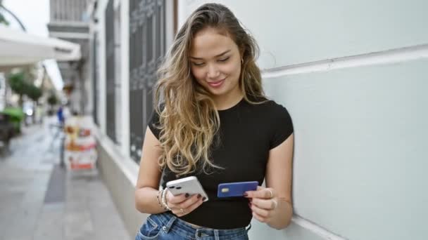 Genç ve güzel İspanyol bir kadın akıllı telefon ve kredi kartıyla sokakta alışveriş yapıyor. - Video, Çekim