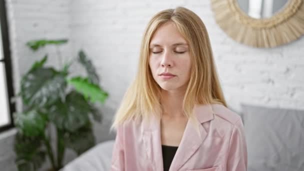 Una mujer rubia tranquila medita en un dormitorio elegante con una expresión serena y las manos en posición de oración. - Metraje, vídeo