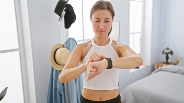 Eine fokussierte Frau überprüft ihren Fitness-Tracker in einem hellen Schlafzimmer, was auf einen modernen Lebensstil und ein modernes Gesundheitsbewusstsein hindeutet. - Filmmaterial, Video