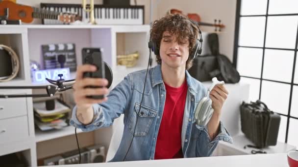 Jonge volwassen man met hoofdtelefoon neemt een selfie in een muziekstudio, presentatie van technologie en entertainment. - Video