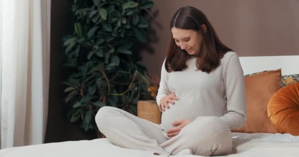 Těhotenství Glow koncept brzy-budoucí máma sedí na posteli, její krátké vlasy rámují její šťastný obličej, zatímco ona hladí její rostoucí břicho s mírumilovným úsměvem. - Záběry, video