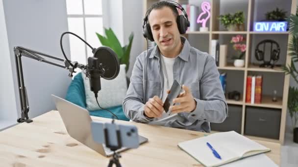 Mosolygó ember fejhallgatóval egy podcast stúdióban kölcsönhatásban okostelefonnal és felvevő berendezés - Felvétel, videó
