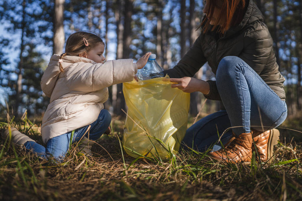 Mutter und Tochter kleine kaukasische Mädchen Kleinkind und Frau sammeln Plastikflaschen Müll aus dem Wald Naturpark steckte es in die Tasche - Foto, Bild