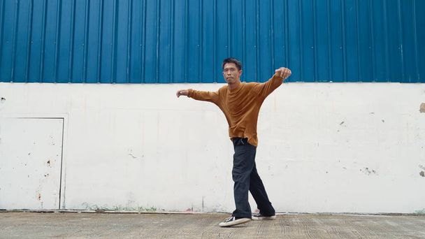 Hispanic man stretch arms and dance street dancing in front of wall. Motion shot of stylish dancer or choreographer in casual outfit practicing dancing in hip hop style. Outdoor sport 2024. Endeavor. - Photo, Image