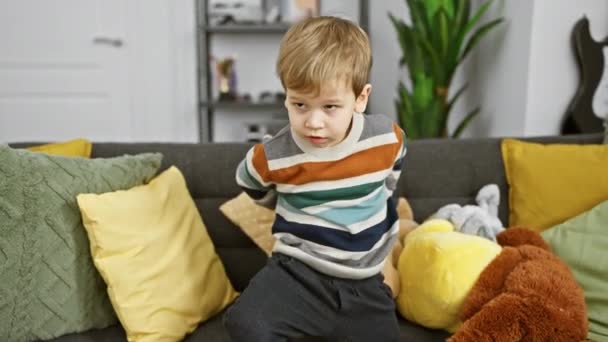 Um menino de criança branca loira sentado em um sofá cinza com travesseiros coloridos e brinquedos de pelúcia em uma sala de estar aconchegante. - Filmagem, Vídeo