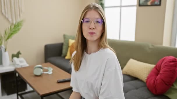 A beautiful, blonde woman wearing glasses poses playfully indoors in a tastefully decorated living room - Footage, Video