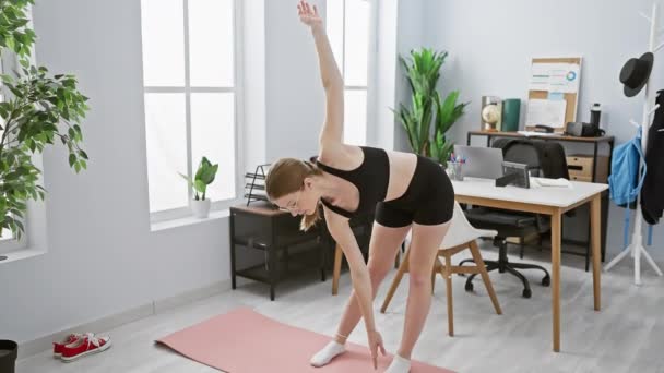 Młoda kobieta rozciąga się na macie do jogi w nowoczesnym biurze, prezentując fitness i wellness w miejscu pracy. - Materiał filmowy, wideo