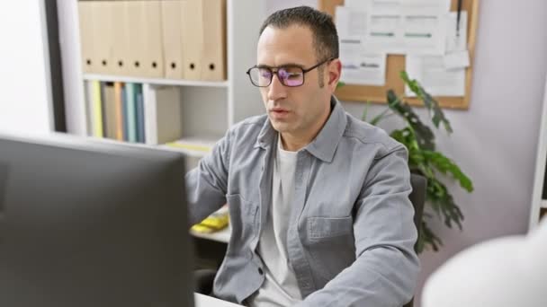 Um jovem hispânico com uma camisa cinza sentindo-se cansado em sua mesa de escritório, tirando os óculos com um computador e plantas de interior no fundo. - Filmagem, Vídeo