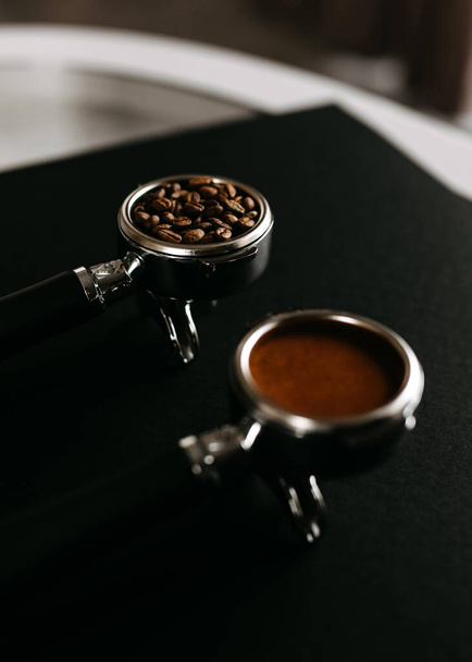 Two espresso portfilters on a black surface, one filled with coffee beans and the other with ground coffee. - Фото, изображение