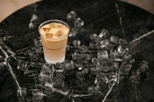 Un café helado en una taza de plástico rodeado de cubitos de hielo dispersos en una superficie de mármol. - Foto, imagen