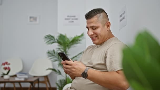 Fröhlicher junger Mann aus Lateinamerika, der fröhlich auf seinem Smartphone SMS schreibt, während er gemütlich im Wartezimmerstuhl sitzt - Filmmaterial, Video