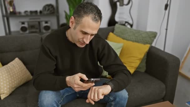 Um homem focado verifica o tempo em seu smartwatch enquanto segura um telefone em uma sala de estar aconchegante. - Filmagem, Vídeo
