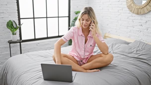 Una joven rubia en pijama trabaja en un portátil y habla por teléfono mientras está sentada con las piernas cruzadas en una cama en un dormitorio luminoso. - Imágenes, Vídeo
