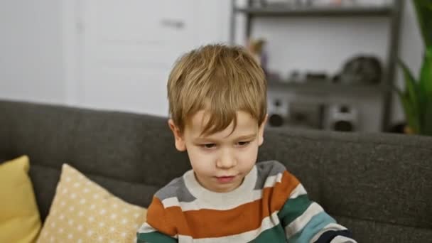 Niño caucásico con el pelo rubio se sienta en el interior de un sofá gris, rodeado de almohadas de colores en una acogedora sala de estar. - Metraje, vídeo