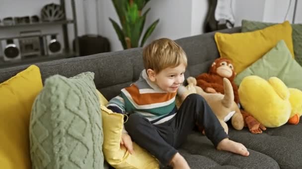 Egy játékos kisgyerek mosolyog egy kényelmes kanapén plüss játékokkal egy színes nappaliban.. - Felvétel, videó