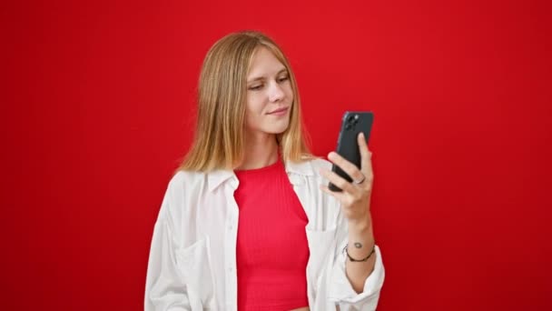 Blond vrouw nemen selfie met smartphone tegen rode achtergrond, uiten vreugde en het maken van vrede teken. - Video