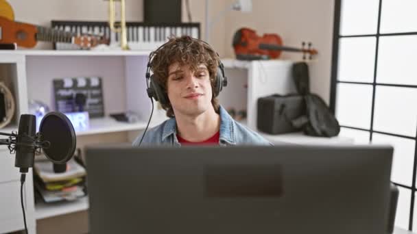 Przystojny mężczyzna z kręconymi włosami nagrywający muzykę w nowoczesnym studiu, noszący słuchawki przed mikrofonem i laptopem - Materiał filmowy, wideo