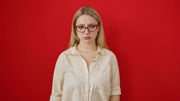 Depressziós, fiatal szőke, ideges arckifejezéssel, dühében és félelmében sír. szemüveget visel, álló elszigetelt felett piros háttér - Felvétel, videó