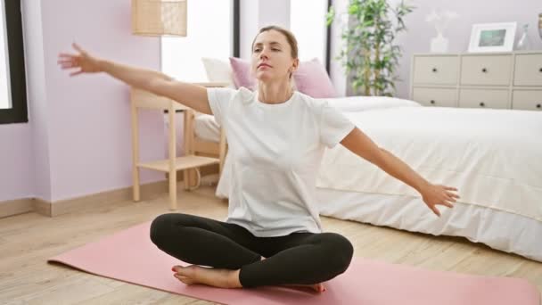 Une jeune femme caucasienne s'étire sur un tapis de yoga dans une chambre soigneusement organisée avec des tons blancs et violets, respirant calme et sérénité. - Séquence, vidéo