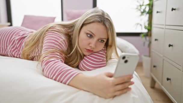 Mietteliäs nuori nainen makaa sängyllään selaamassa älypuhelintaan viihtyisässä makuuhuoneessa - Materiaali, video