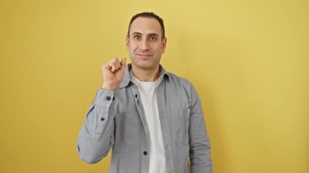 Cheerful young hispanic male in shirt, confidently pointing upward with number one finger, radiating positivity on isolated yellow wall background - Footage, Video