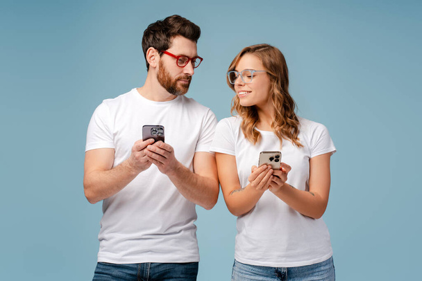 Zajímavý pár při pohledu na smartphony jeden druhého izolované na modrém pozadí. Šťastní přátelé držící mobilní telefony, komunikace online. Technologie, koncept závislosti na sociálních médiích - Fotografie, Obrázek