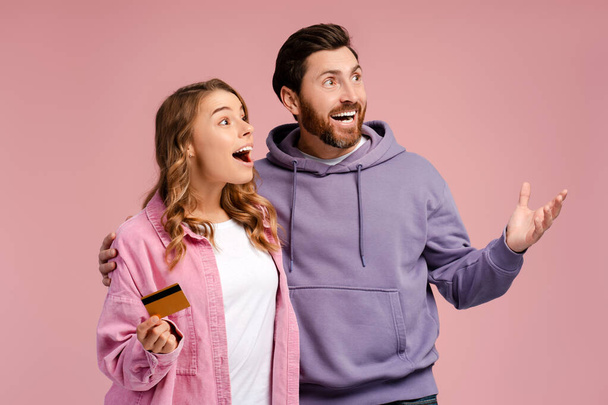 Couple souriant tenant la carte de crédit et se sentant heureux, faire du shopping en ligne isolé sur fond rose. Heureux homme et femme étreignant, se tenant ensemble, détournant les yeux, copiant l'espace. Concept technologique - Photo, image