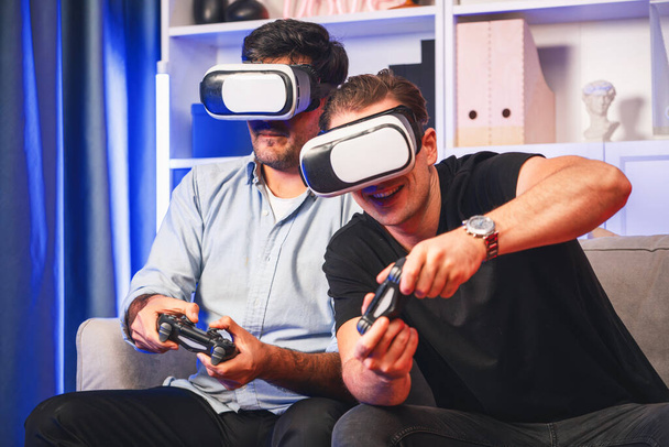 Φιλαράκια παίκτες που παίζουν βιντεοπαιχνίδια χρησιμοποιώντας joysticks και VR ακουστικά εικονικής τεχνολογίας στην πραγματικότητα στο στούντιο με νέον μπλε φως. Comfy ζουν σε εσωτερικούς χώρους με χαρούμενα νικητής πάλης. Πωλητέα. - Φωτογραφία, εικόνα
