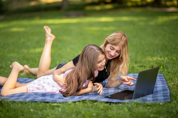 Μητέρα και κόρη κοιτάζουν χαρούμενα ένα λάπτοπ στο πάρκο.. - Φωτογραφία, εικόνα