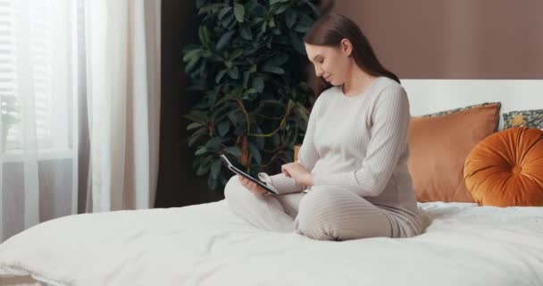 Ελκυστική νεαρή έγκυος γυναίκα κάθεται στο λωτό θέτουν, ατενίζοντας τις φωτογραφίες στο tablet της, ενώ αγκαλιάζει χτύπημα του μωρού της με ένα ήρεμο χαμόγελο. - Πλάνα, βίντεο