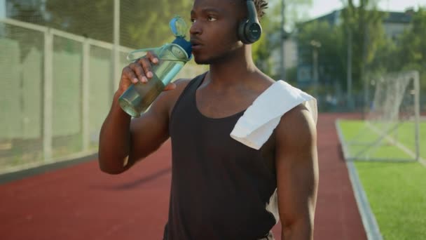Африканський американський спортивний чоловік в навушниках слухає музику втомлений спраглий спортсмен бігун п'є воду здорового ранкового зволоження після тренування на міському стадіоні спортсмен п'є зі спортивної пляшки на відкритому повітрі - Кадри, відео