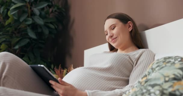 Dreamy alluring pregnant woman dressed in a stylish and comfortable outfit, sitting on a bed, holding a tablet, and admiring photos with interest. - Footage, Video
