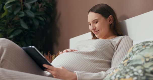 Delighted pregnant woman in a trendy and comfortable outfit sits on a bed, holding a tablet, and browsing through photos with a smile on her face. - Footage, Video