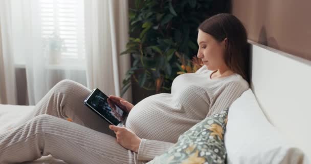 Attractive pregnant woman in a stylish and comfortable outfit sits on a bed holding a tablet, scrolling through photos with a curious expression. - Footage, Video