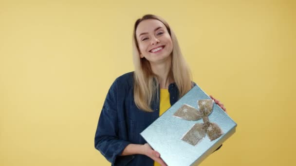 Porträt einer charmanten jungen kaukasischen Frau, die eine schöne grüne Geschenkschachtel mit Goldschleife präsentiert, während sie über gelbem Studiohintergrund posiert. Konzept von Überraschung, Urlaub, Feier und Menschen. - Filmmaterial, Video