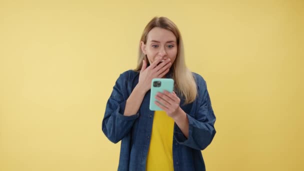 Nuori onnellinen kaukasialainen nainen osoittaa matkapuhelinta eristettynä keltaisella taustalla. Hymyilevä nainen, jolla on gadget ja joka ilmaisee positiivisia tunteita sisätiloissa. Voiton ja voiton käsite. - Materiaali, video