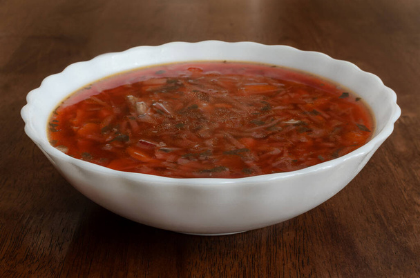 Na zdjęciu biała ceramiczna miska zupy pomidorowej umieszczona na drewnianej powierzchni. Zupa jest czerwona i wydaje się gruba, z widocznymi kawałkami warzyw i ziół. - Zdjęcie, obraz