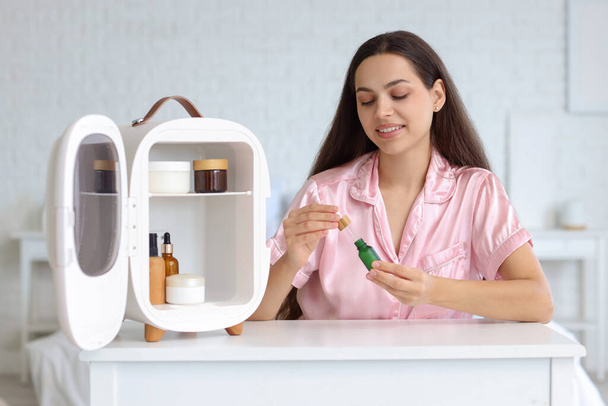 Hermosa joven feliz mujer con botella de aceite y nevera pequeña para productos cosméticos en la mesa en el dormitorio - Foto, imagen