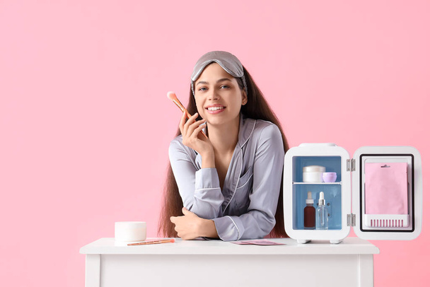 Beautiful young happy woman with brush and small refrigerator for cosmetic products on table against pink background - Photo, Image