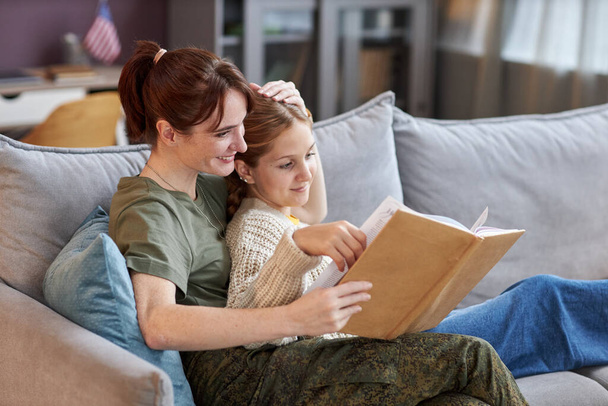 Retrato de vista lateral de mujer militar con hija en casa leyendo libro juntos y relajándose en el sofá - Foto, Imagen