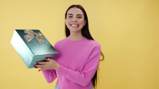Kobieta w różowych ubraniach trzyma błyszczące pudełko z kokardą i uśmiechem stojąc w studio z żółtym tłem. Młoda kobieta z długimi włosami pokazując prezent urodzinowy i wyrażając zachwyt. - Materiał filmowy, wideo